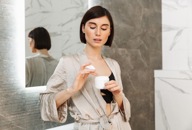 Foto de mulher bonita morena em pé perto do espelho e segurando o banco com creme para o rosto em casa banheiro