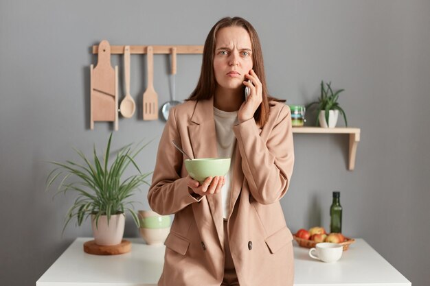 Foto de mulher de cabelos castanhos triste e atônita vestindo terno bege em pé perto da mesa na cozinha em casa tomando café da manhã ou jantando segurando o prato falando no celular ouvindo notícias chocadas