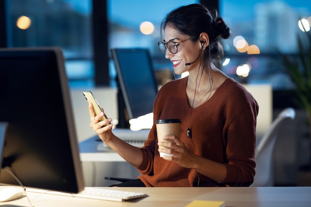 Foto de mulher de negócios jovem bonita tendo videocall com telefone celular enquanto trabalhava com o computador sentado no escritório.