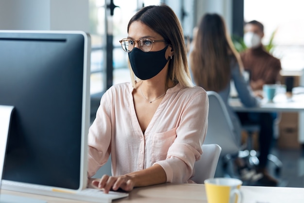 Foto de mulher de negócios jovem bonita usando uma máscara higiênica, trabalhando com o computador enquanto está sentado na mesa no escritório moderno de inicialização.