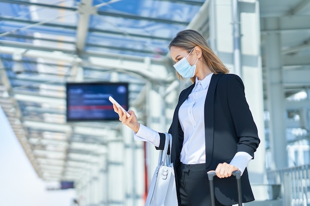 Foto de mulher elegante usando máscara de proteção andando com bolsa e mala na estação ferroviária