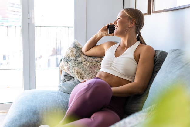 Foto de mulher feliz desportiva falando com seu telefone celular enquanto está sentado no sofá na sala de estar em casa.