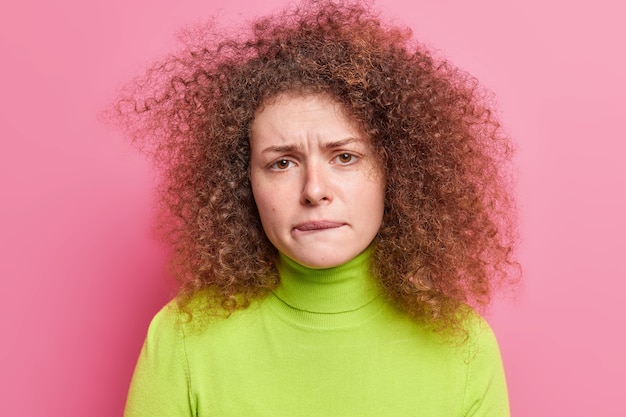 Foto de mulher frustrada e infeliz com cabelo encaracolado morde os lábios parece nervosa e sente-se ansiosa vestida com uma camiseta verde casual isolada sobre a parede rosa. Mulher perplexa e perturbada