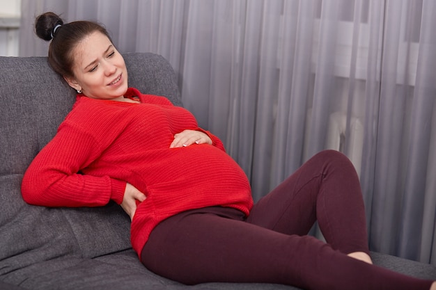 Foto de mulher grávida insatisfeita sente dor nas costas, cara carrancuda de descontentamento, veste roupas casuais, tem problemas com a saúde, senta-se no sofá confortável.