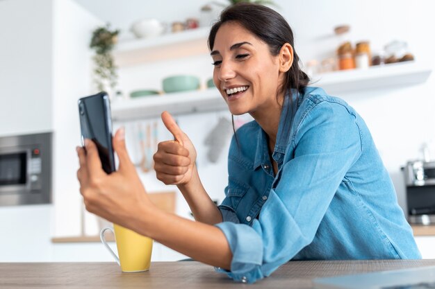 Foto de mulher jovem e bonita fazendo videochamada e mostrando o polegar para cima para smartphone durante o café da manhã em casa.