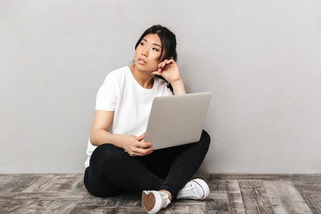 Foto de mulher jovem e bonita pensando asiático posando isolado sobre uma parede cinza, usando o computador portátil.