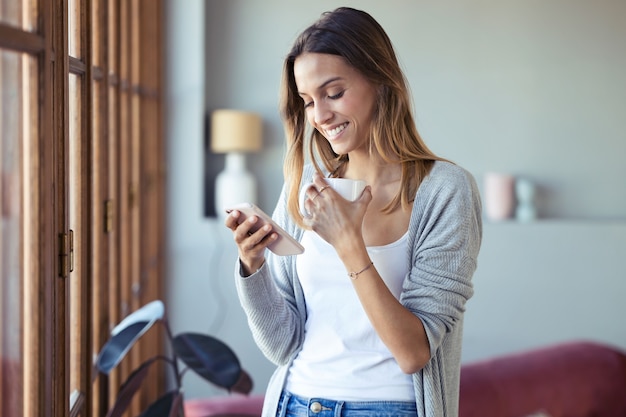 Foto foto de mulher jovem e bonita usando seu telefone celular enquanto bebia café perto da janela da sala de estar em casa.