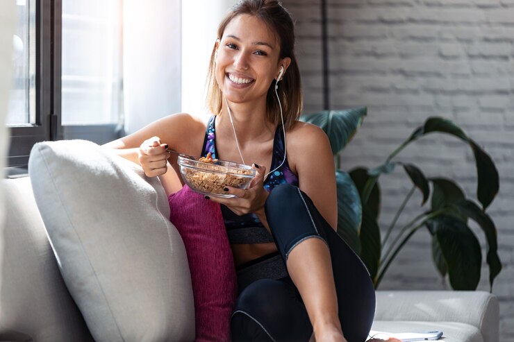 Foto foto de mulher jovem e desportiva comendo uma tigela de muesli enquanto ouve música, sentado no sofá em casa.