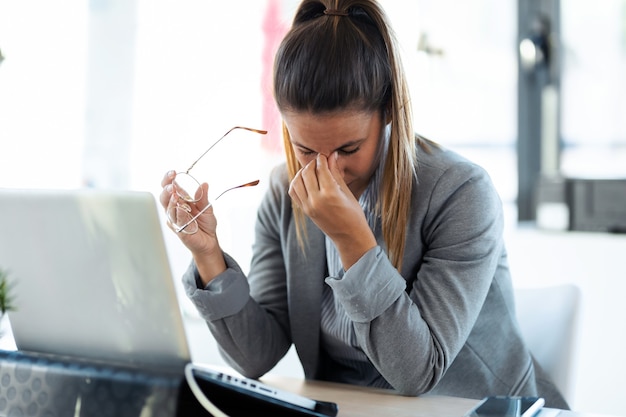 Foto de mulher jovem exausta com dor de cabeça, trabalhando com seu laptop no escritório.