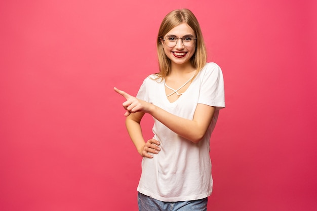 Foto de mulher jovem feliz em pé isolada sobre fundo rosa. olhando a câmera mostrando o apontar do copyspace.