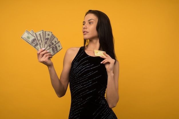 Foto de mulher jovem feliz satisfeito posando isolado sobre o fundo da parede amarela, segurando dinheiro e cartão de crédito ou dívida.