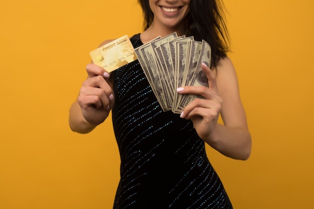 Foto de mulher jovem feliz satisfeito posando isolado sobre o fundo da parede amarela, segurando dinheiro e cartão de crédito ou dívida.