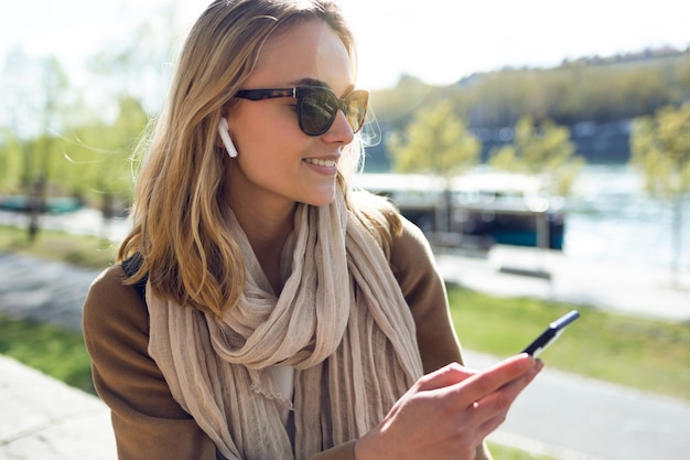 Foto de mulher jovem, ouvindo música com fones de ouvido sem fio e o smartphone na rua.