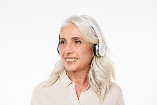 Foto de mulher madura dos anos 60 com cabelos grisalhos, sorrindo com dentes perfeitos e olhando de lado enquanto ouve música através de fones de ouvido sem fio, isolados sobre a parede branca