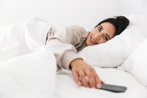 Foto de mulher morena de 30 anos usando smartphone, deitada na cama com lençóis brancos em casa