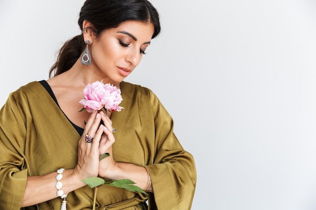 Foto de mulher morena indiana com maquiagem, usando joias étnicas e vestido de seda tradicional, posando com uma flor isolada sobre uma parede branca