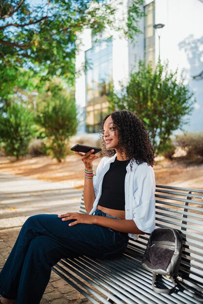 Foto de mulher negra enviando uma mensagem de voz para amigos enquanto ela está sentada em um banco no parque