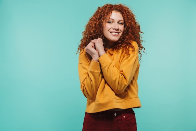 Foto de mulher positiva de 20 anos com cabelo ruivo cacheado sorrindo e olhando para a câmera isolada sobre a parede azul