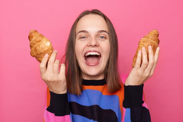 Foto de mulher rindo animada com cabelo castanho vestindo suéter segurando dois croissants nas mãos gritando sente fome posando isolado sobre fundo rosa