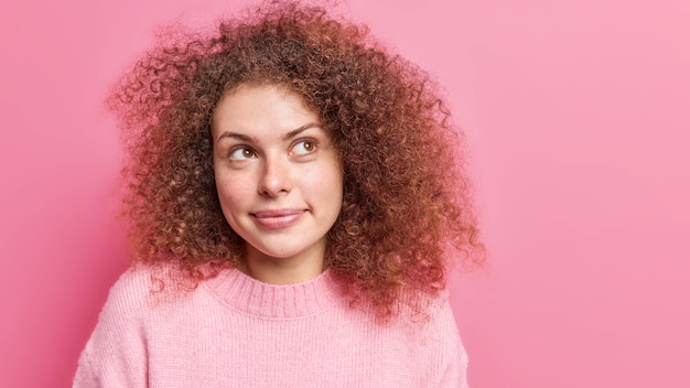 Foto de mulher saudável com cabelo crespo e encaracolado parece pensativamente de lado, concentrada acima de estar imersa em pensamentos, usa um macacão básico isolado na parede rosa com espaço em branco para seu texto