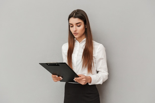 Foto de mulher séria de camisa branca e saia preta segurando uma prancheta com documentos no escritório, isolada sobre uma parede cinza