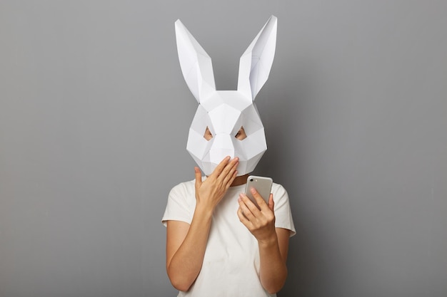 Foto de mulher surpresa chocada vestindo camiseta branca e máscara de coelho de papel em pé isolada sobre fundo cinza usando telefone celular lendo notícias de última hora cobrindo a boca com a palma da mão