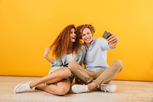 Foto de namorado e namorada alegres sentados no chão juntos e tirando uma selfie no smartphone, isolada sobre fundo amarelo