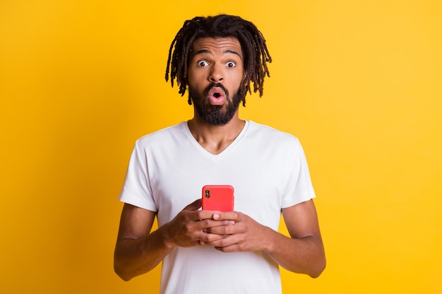 Foto de negro segurando telefone olhar câmera expressão chocada vestir camiseta branca isolado fundo de cor amarela