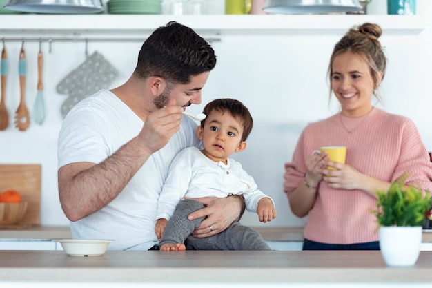 Foto de pai jovem bonito alimentando seu filho bebê enquanto a mãe os procurava na cozinha em casa.