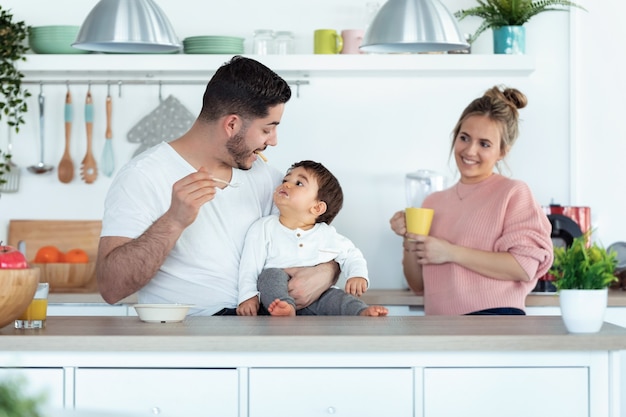 Foto de pai jovem bonito alimentando seu filho bebê enquanto a mãe os procurava na cozinha em casa.