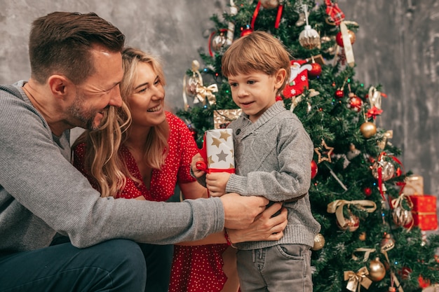 Foto de papai, mãe e filho. Passar tempo juntos. Árvore de Natal em casa, momentos felizes. Preparando-se para as férias. Família com um filho. Felizes juntos. Divirta-se com a caixa de presente. Cores verdes REd