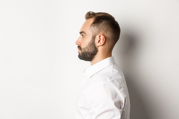 Foto de perfil de close-up de um homem barbudo bonito, olhando para a esquerda, em pé contra um fundo branco.