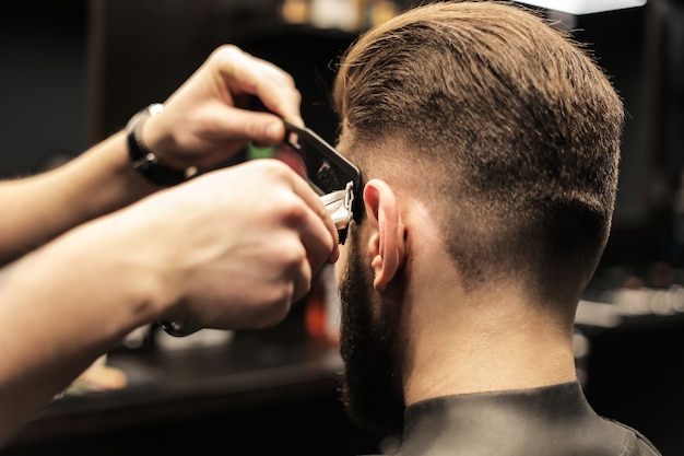 Foto foto de perfil de um jovem barbeiro aparando o cabelo de seu cliente com um barbeador elétrico e um pente em uma barbearia.