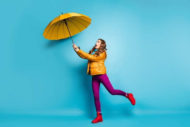 Foto de perfil em tamanho real de uma senhora bonita viajante segurando guarda-chuva brilhante voando com o vento soprando usar sobretudo amarelo lenço calça violeta calçado vermelho parede de cor azul isolada