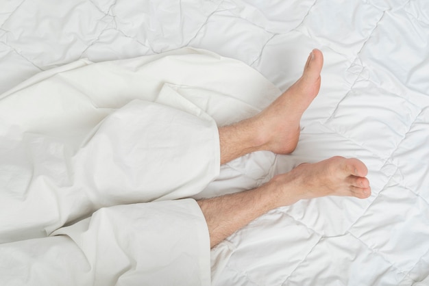 Foto de pés masculinos na cama, debaixo do lençol. Vista do topo. Homem dormindo na cama, debaixo do cobertor.