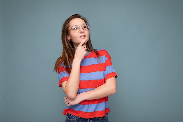 Foto de positiva pensativa jovem bonita morena com emoções sinceras, vestindo camiseta listrada casual e óculos ópticos, isolados sobre um fundo azul com espaço de cópia.