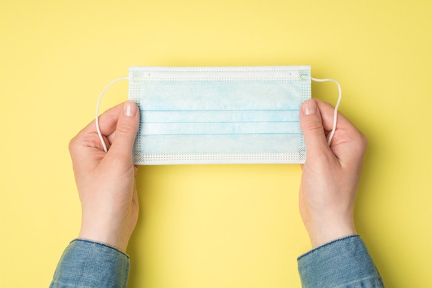 Foto de primeira pessoa da vista superior da mão de uma mulher segurando a máscara médica em fundo amarelo isolado com espaço em branco