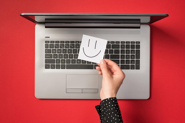 Foto de primeira pessoa da vista superior da mão segurando o papel de nota adesivo branco com rosto sorridente desenhado sobre laptop cinza aberto em fundo vermelho isolado
