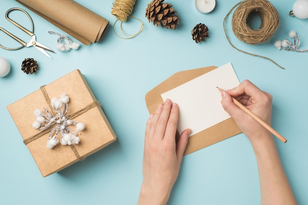 Foto de primeira pessoa da vista superior das mãos escrevendo envelope de carta caixa de presente de papel kraft com galho de neve e guita bolas de árvore de natal ferramentas de artesanato em fundo azul pastel isolado com espaço em branco