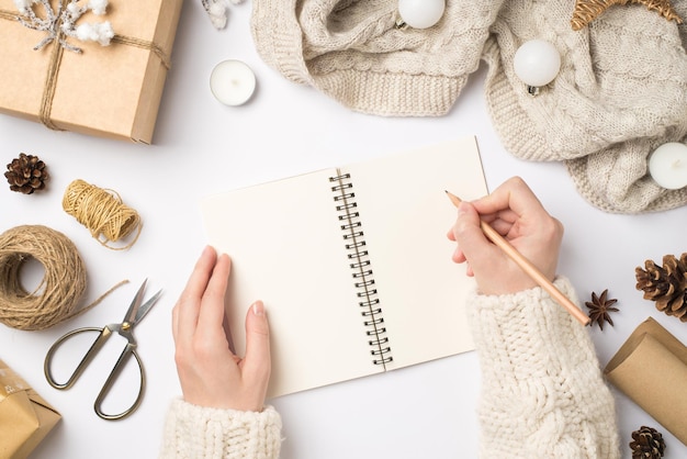 Foto de primeira pessoa da vista superior das mãos no suéter escrevendo com lápis no planner giftbox cachecol bolas de árvore de natal e ferramentas de artesanato em fundo branco isolado com espaço vazio