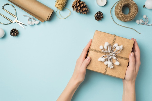 Foto de primeira pessoa de vista superior de mãos segurando uma caixa de presente de papel kraft decorada com galho de neve e ferramentas de artesanato de bolas de árvore de natal branca em fundo azul pastel isolado com espaço vazio