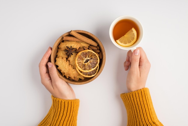 Foto de primeira pessoa vista superior das mãos femininas na camisola amarela segurando a xícara de chá e tigela de madeira com biscoitos fatias de limão secas anis e paus de canela em fundo branco isolado