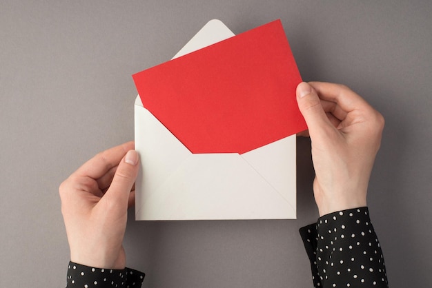 Foto de primeira pessoa vista superior de mãos femininas segurando um envelope branco aberto com cartão vermelho em fundo cinza isolado com copyspace