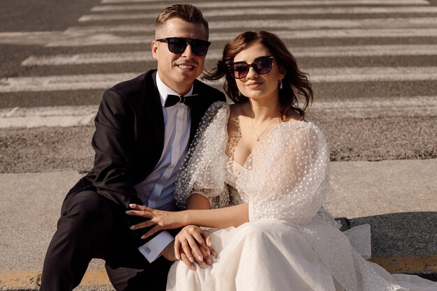 Foto de recém-casados apaixonados, a noiva e o noivo em vestido de noiva estão sentados na faixa de pedestres, num contexto de paisagem urbana, conceito de casamento, óculos de sol nos olhos.