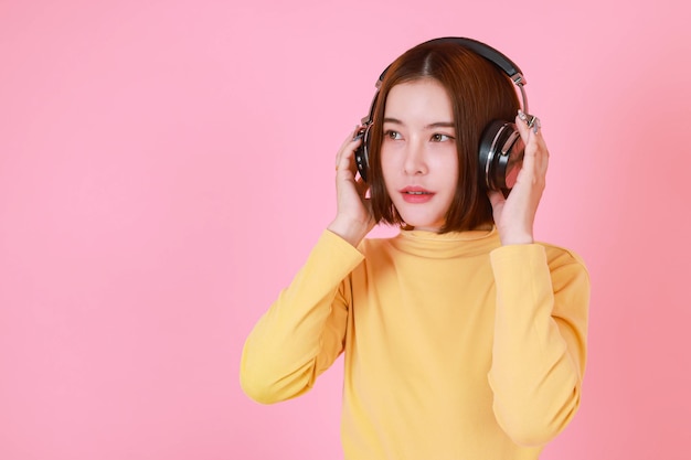 Foto de recorte de estúdio de retrato de modelo feminino de cabelo curto bonito jovem asiático em pé de camisa de manga longa amarela segurando o ajuste de fones de ouvido pretos grandes enquanto ouve a música no fundo rosa.