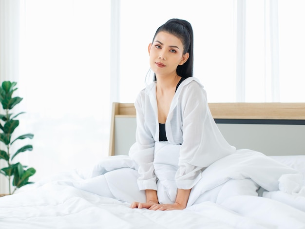 Foto de retrato da mulher sexy asiática do milênio em regata de conforto casual e camisa sentada sob um cobertor quente branco grosso posando esticando o braço depois de acordar no quarto de manhã em casa