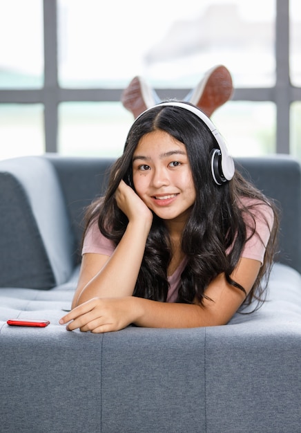 Foto de retrato de jovem adolescente relaxado deitado no sofá cinza, ouvindo música com fone de ouvido e jogando o telefone celular. Jovem júnior segurando o smartphone e olhando para a câmera