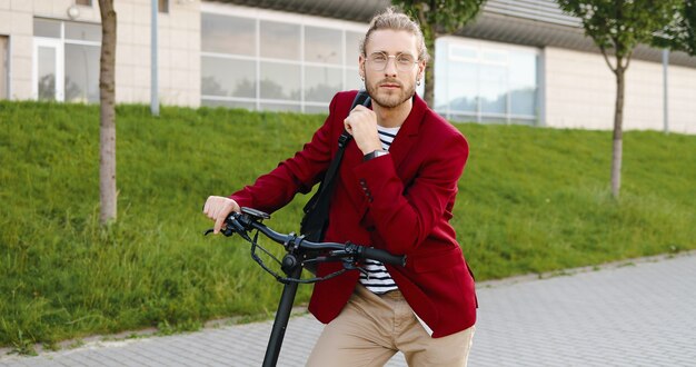 Foto de retrato de jovem bonito caucasiano com casaco vermelho e óculos em pé ao ar livre com scooter elétrico e olhando para a câmera. Cara elegante em veículo na rua da cidade. Homem bonito posando.