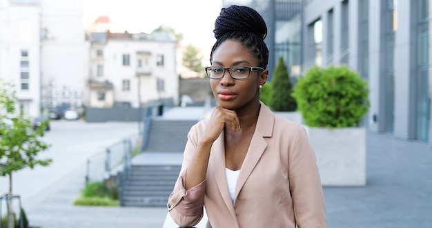 Foto de retrato de uma jovem mulher afro-americana bonita de óculos, olhando para a câmera ao ar livre e inclinando-se com o queixo na mão. mulher de negócios séria confiante atraente na rua perto do centro de negócios.