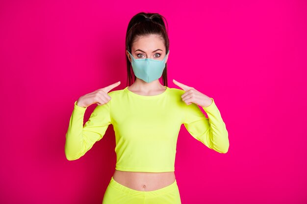 Foto de retrato de uma menina apontando para uma máscara protetora médica usando um rabo de cavalo esportivo isolado em um fundo de cor fúcsia vibrante